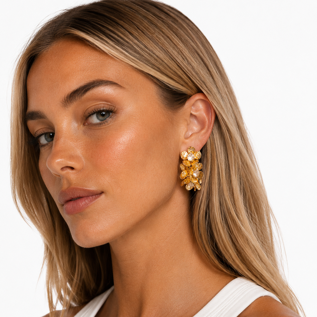 A close-up of a woman with blonde hair wearing a white ribbed tank top and a gold floral cluster earring, set against a clean white background.