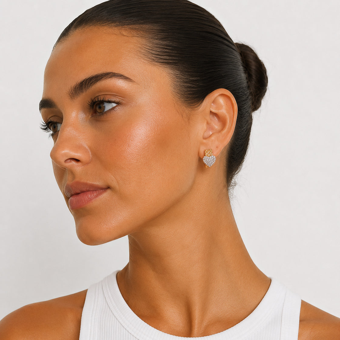 A close-up of a woman with her hair in a bun wearing a small heart-shaped stud earring with a sparkling clear stone setting, paired with a white ribbed tank top against a light gray background.