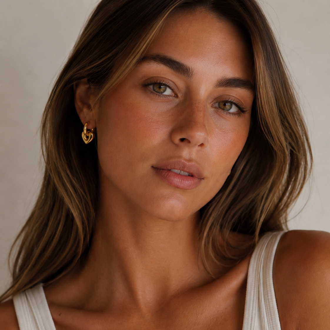 Woman wearing gold heart huggie earrings on a neutral background